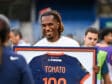 Smiling man with dreadlocks holding a framed jersey displaying the number 100 at an event.