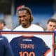Smiling man with dreadlocks holding a framed jersey displaying the number 100 at an event.