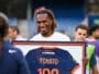Smiling man with dreadlocks holding a framed jersey displaying the number 100 at an event.