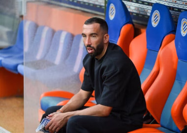 Man seated on a team bench in a stadium, wearing a black shirt and looking focused.