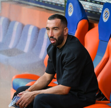 Man seated on a team bench in a stadium, wearing a black shirt and looking focused.