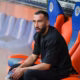 Man seated on a team bench in a stadium, wearing a black shirt and looking focused.