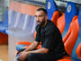 Man seated on a team bench in a stadium, wearing a black shirt and looking focused.
