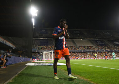 Montpellier Herault SC v Dijon FCO - Ligue 2 BKT 20262027 | AllezPaillade.com Soccer player in a blue and orange jersey number 19 standing near the sideline during a night match, stadium filled with spectators in the background.