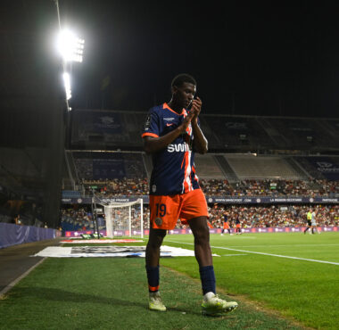 Soccer player in a blue and orange jersey number 19 standing near the sideline during a night match, stadium filled with spectators in the background.
