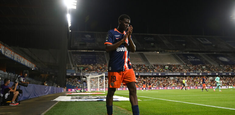 Soccer player in a blue and orange jersey number 19 standing near the sideline during a night match, stadium filled with spectators in the background.