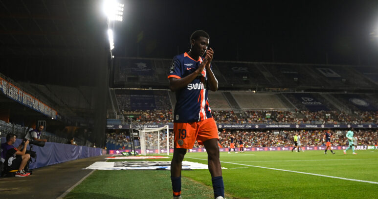 Soccer player in a blue and orange jersey number 19 standing near the sideline during a night match, stadium filled with spectators in the background.