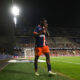 Soccer player in a blue and orange jersey number 19 standing near the sideline during a night match, stadium filled with spectators in the background.