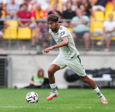 Soccer player in a white kit dribbling the ball on a green pitch with a blurred crowd in yellow seats behind him.