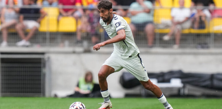 Soccer player in a white kit dribbling the ball on a green pitch with a blurred crowd in yellow seats behind him.