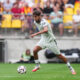 Soccer player in a white kit dribbling the ball on a green pitch with a blurred crowd in yellow seats behind him.
