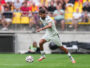 Soccer player in a white kit dribbling the ball on a green pitch with a blurred crowd in yellow seats behind him.