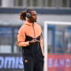 Athlete wearing a peach and black tracksuit with over-ear headphones, holding a phone on a sports field surface.