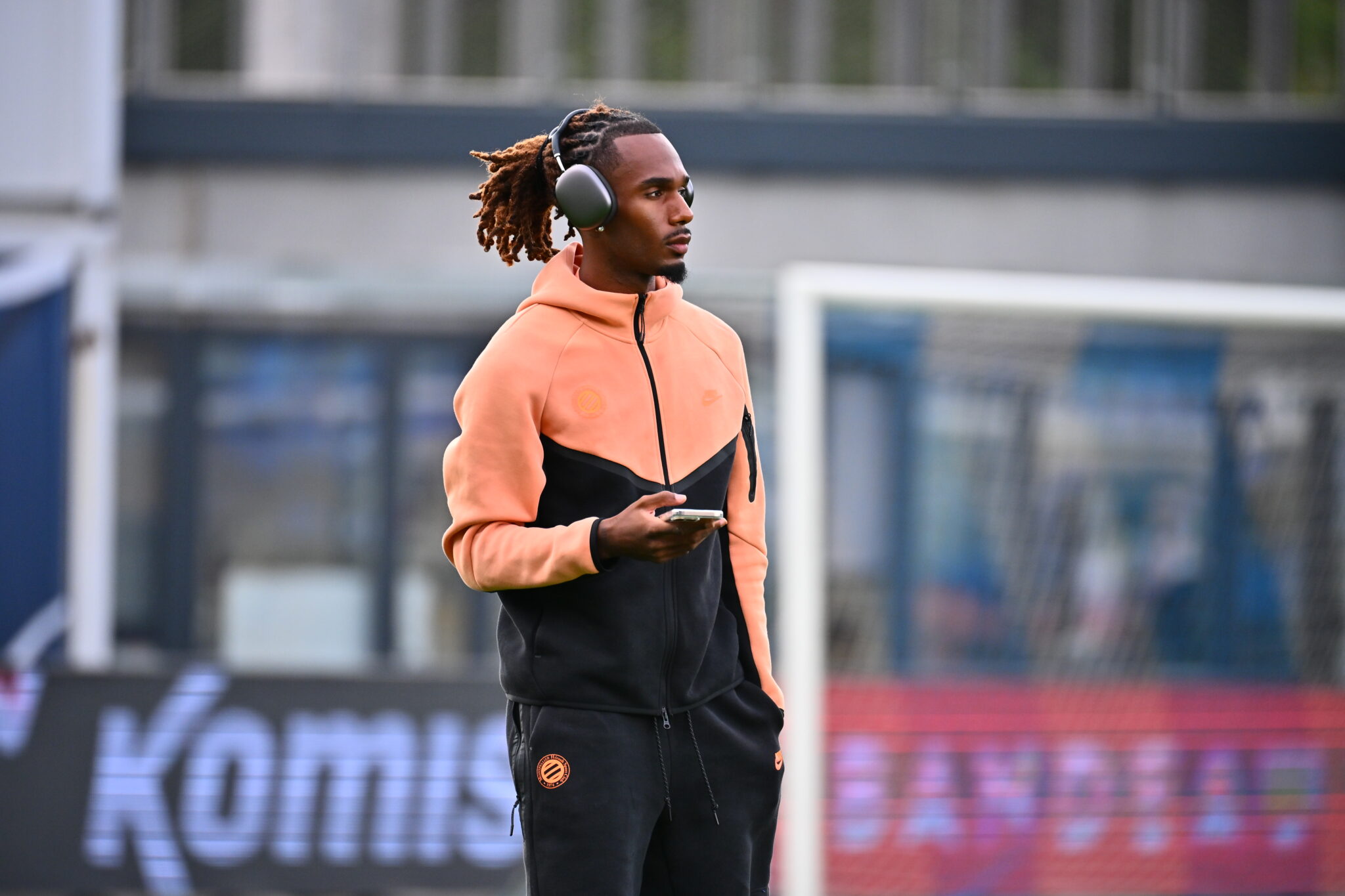 Athlete wearing a peach and black tracksuit with over-ear headphones, holding a phone on a sports field surface.