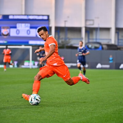 Soccer player in an orange kit dribbles the ball on a green pitch with a blurred stadium and scoreboard in the background.