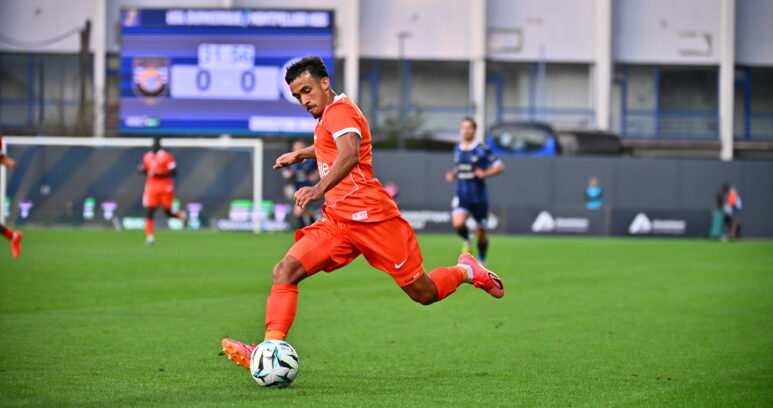 Soccer player in an orange kit dribbles the ball on a green pitch with a blurred stadium and scoreboard in the background.