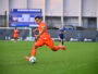 Soccer player in an orange kit dribbles the ball on a green pitch with a blurred stadium and scoreboard in the background.
