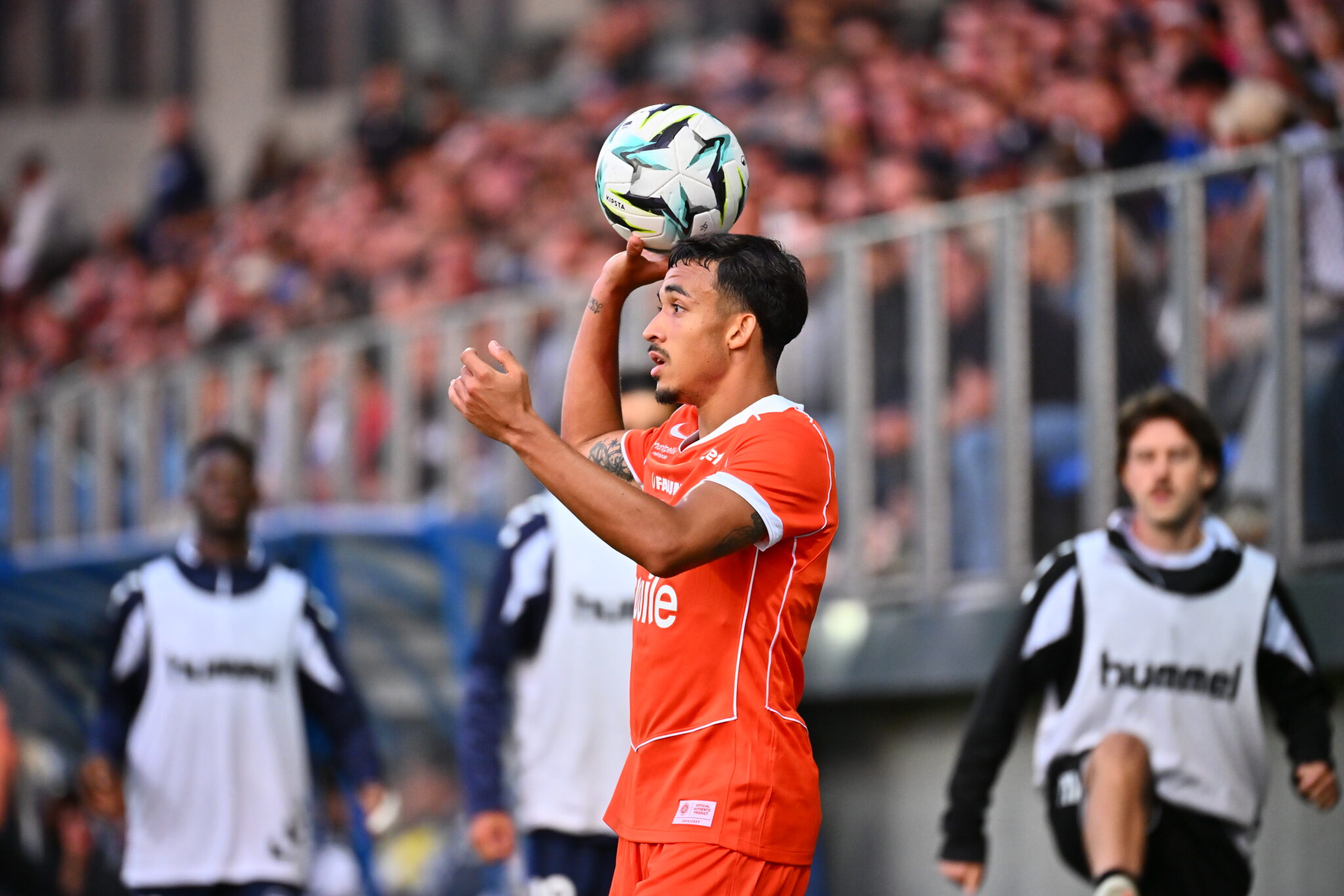 Soccer player in an orange jersey heads a soccer ball during a match, with a blurred crowd in the stands behind him on the field behind.
