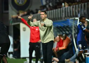 Football coach on the sideline gesturing with both hands during a match, crowd in stands behind him.