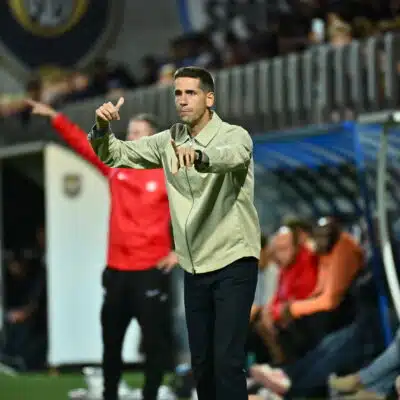 Football coach on the sideline gesturing with both hands during a match, crowd in stands behind him.