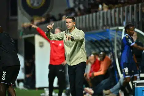 Football coach on the sideline gesturing with both hands during a match, crowd in stands behind him.
