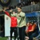 Football coach on the sideline gesturing with both hands during a match, crowd in stands behind him.