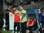Football coach on the sideline gesturing with both hands during a match, crowd in stands behind him.