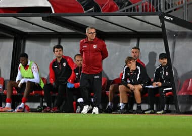 Soccer team on the bench during a match, coach standing and looking ahead while players sit in red-and-black gear under a dugout roof on the sideline.