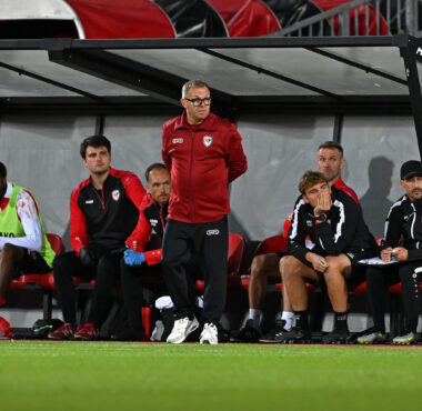 Soccer team on the bench during a match, coach standing and looking ahead while players sit in red-and-black gear under a dugout roof on the sideline.