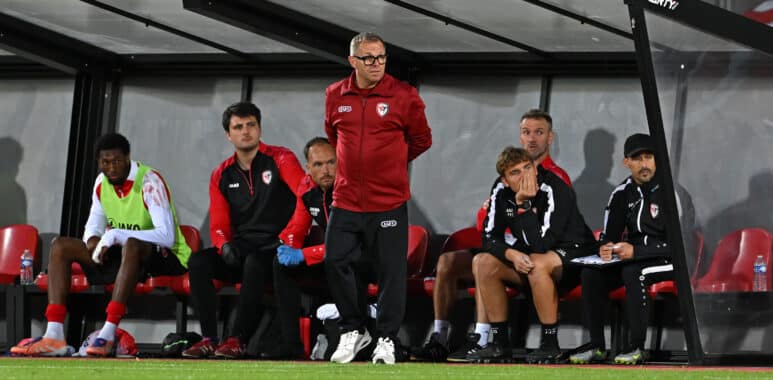 Soccer team on the bench during a match, coach standing and looking ahead while players sit in red-and-black gear under a dugout roof on the sideline.