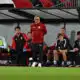Soccer team on the bench during a match, coach standing and looking ahead while players sit in red-and-black gear under a dugout roof on the sideline.