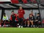 Soccer team on the bench during a match, coach standing and looking ahead while players sit in red-and-black gear under a dugout roof on the sideline.