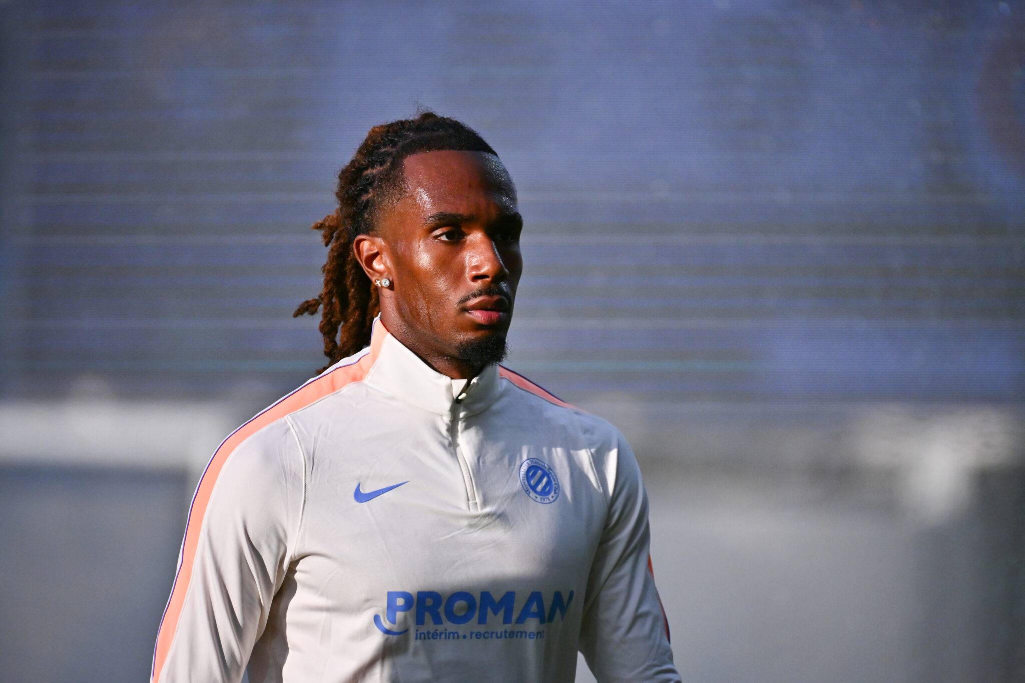 Tchato USL Dunkerque v Montpellier HSC - Ligue 2 BKT 20262027 | AllezPaillade.com Athlete wearing a white training jacket with pink stripes looks off to the side against a blurred stadium backdrop.