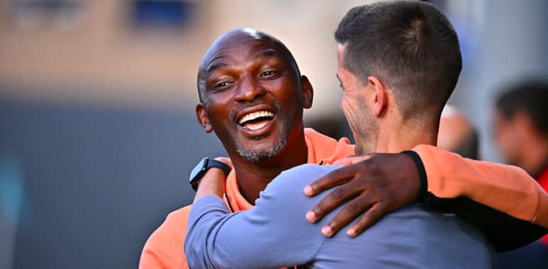 Two men hug warmly and smile at each other outdoors in a friendly greeting.