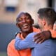 Two men hug warmly and smile at each other outdoors in a friendly greeting.