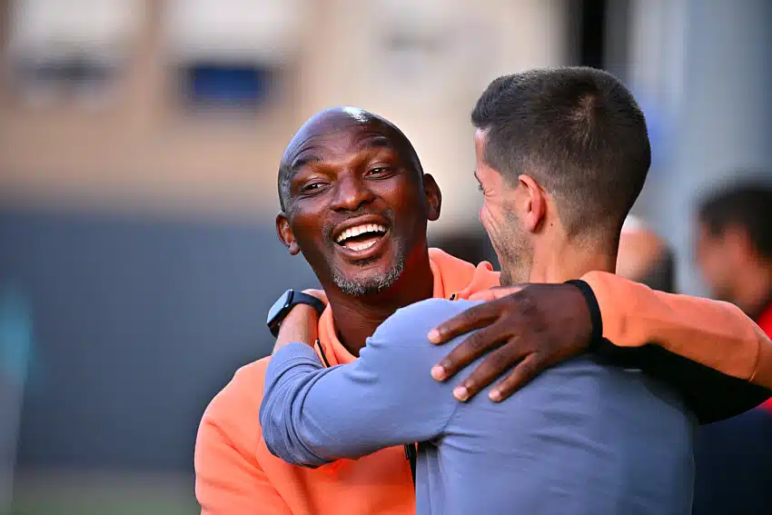 Camara | AllezPaillade.com Two men hug warmly and smile at each other outdoors in a friendly greeting.