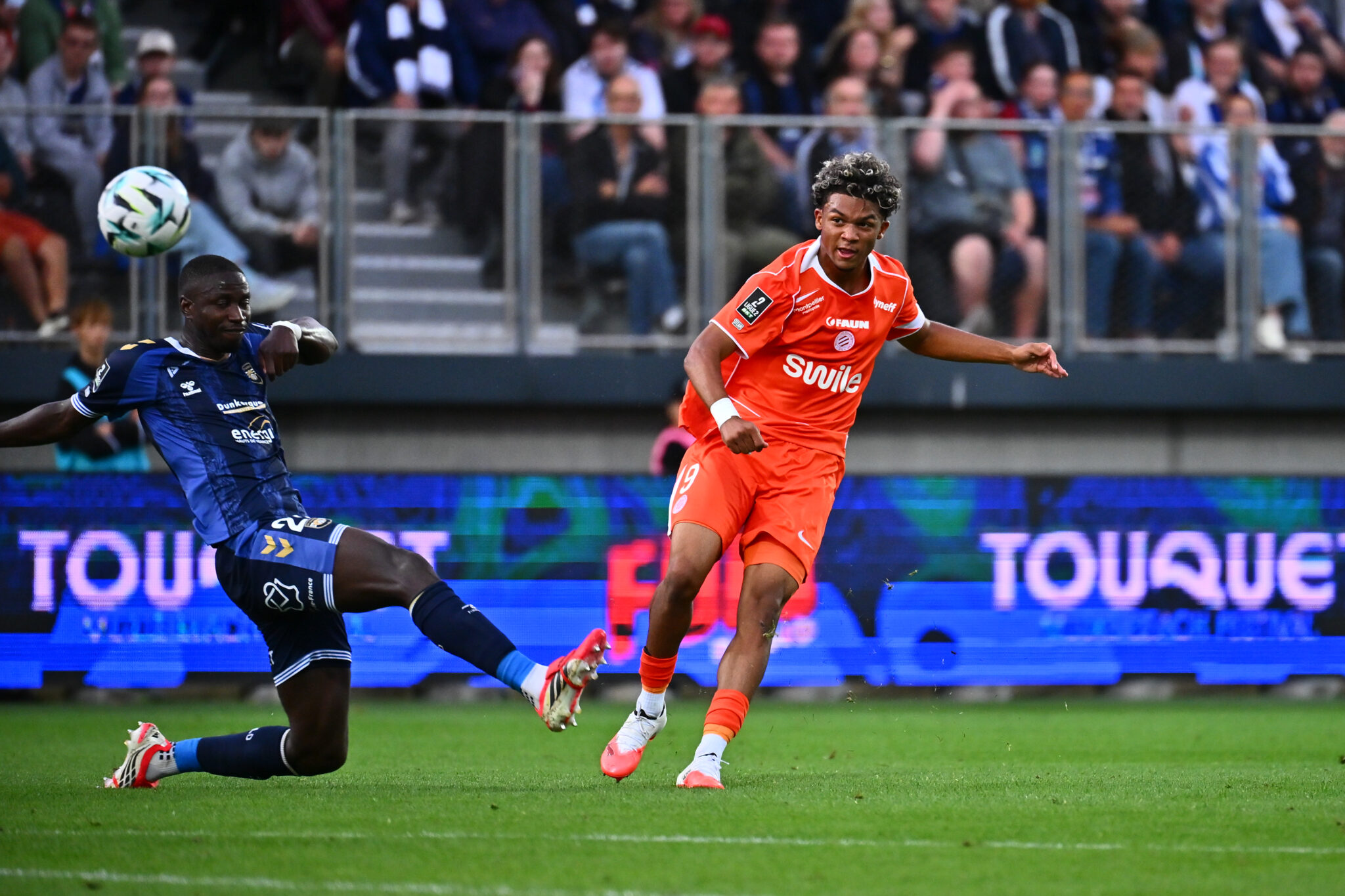 Soccer players clash for the ball on a green pitch; orange kit vs blue kit, with a cheering crowd in the stands behind them.