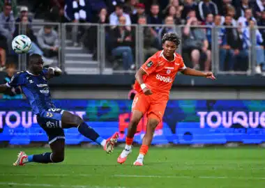 Soccer players clash for the ball on a green pitch; orange kit vs blue kit, with a cheering crowd in the stands behind them.