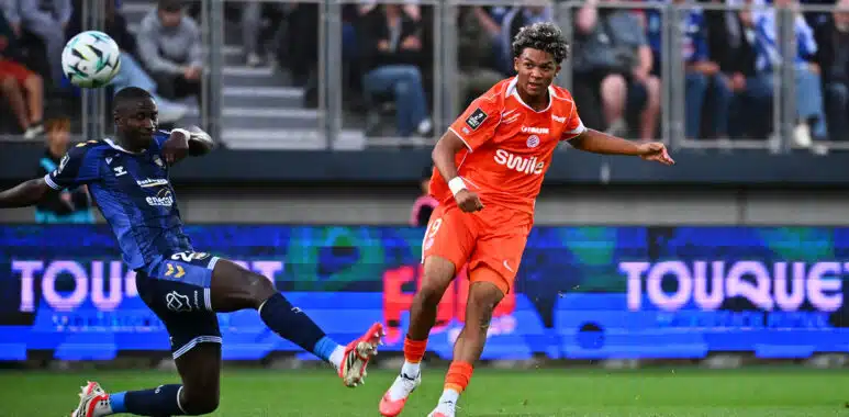 Laciné Megnan USL Dunkerque v Montpellier HSC - Ligue 2 BKT 20262027 | AllezPaillade.com Soccer players clash for the ball on a green pitch; orange kit vs blue kit, with a cheering crowd in the stands behind them.