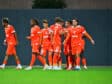 Group of soccer players in bright orange uniforms standing together on a green pitch, celebrating.