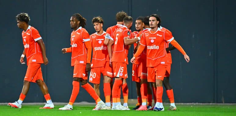 Group of soccer players in bright orange uniforms standing together on a green pitch, celebrating.