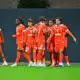Group of soccer players in bright orange uniforms standing together on a green pitch, celebrating.