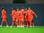 Group of soccer players in bright orange uniforms standing together on a green pitch, celebrating.