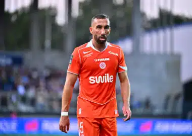 Soccer player in an orange jersey walking on the field with a blurred stadium crowd in the background.