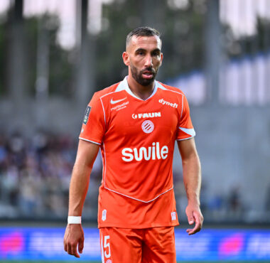 Soccer player in an orange jersey walking on the field with a blurred stadium crowd in the background.