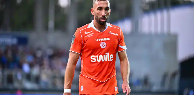 Soccer player in an orange jersey walking on the field with a blurred stadium crowd in the background.