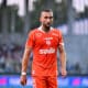 Soccer player in an orange jersey walking on the field with a blurred stadium crowd in the background.