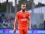 Soccer player in an orange jersey walking on the field with a blurred stadium crowd in the background.