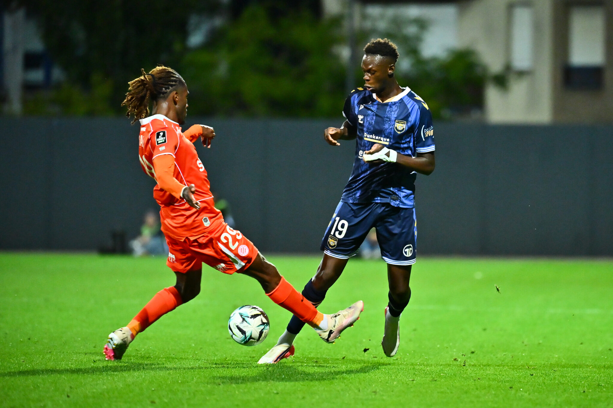 Tchato USL Dunkerque v Montpellier HSC - Ligue 2 BKT 20262027 | AllezPaillade.com Two soccer players compete for the ball on a green field; orange jersey on the left, dark blue jersey on the right, mid-action dribble.
