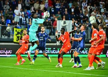 Soccer goalkeeper in teal leaps to catch the ball as players in orange and blue contest in the box with spectators watching.